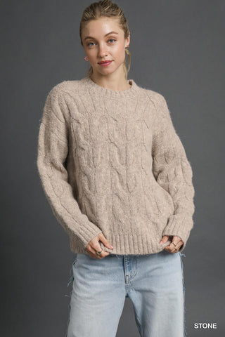 Person wearing a beige cable knit sweater and light blue jeans against a gray background