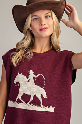 Woman wearing a maroon sleeveless top with a white horse and rider design, holding a brown cowboy hat.