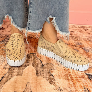 A pair of tan slip-on sneakers with a floral laser-cut design, worn by a person with frayed hem jeans.
