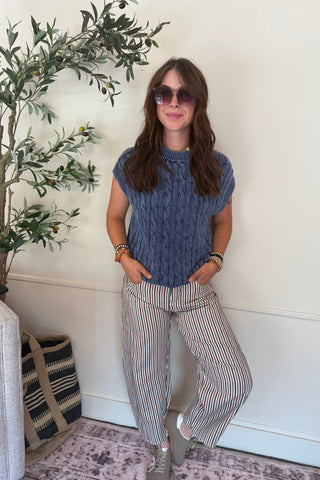 Woman standing indoors next to a plant, wearing a blue sleeveless top and striped pants.