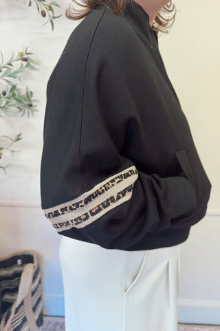 Person wearing a black jacket with patterned cuffs in front of a decorative wall.