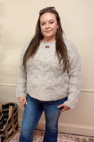 Woman wearing a gray sweater and blue jeans standing in a room with a plant and sofa.