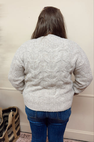 Person wearing a light gray textured sweater and blue jeans standing against a beige wall.