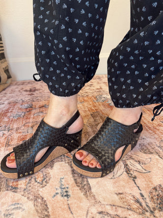 Black woven sandals worn with navy pants on a patterned rug