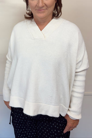 Woman wearing a white sweater standing in front of a decorative wall with greenery.
