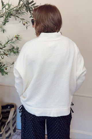 Person wearing a white sweater and dark pants standing near a plant indoors.