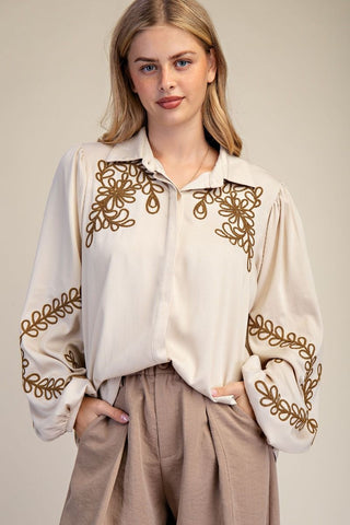 Woman wearing a white blouse with gold embroidery and beige pants on a plain background