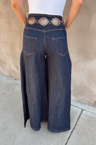 Person wearing wide-leg jeans with a decorative belt against a beige wall.