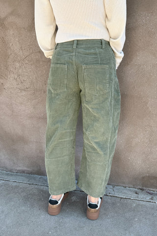 Person wearing green corduroy pants and a light-colored sweater against a brown wall.