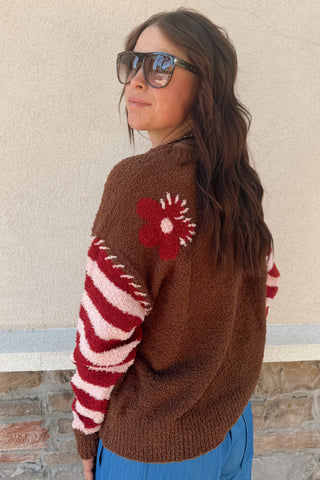 Person wearing a brown sweater with a red flower design, standing against a beige wall.