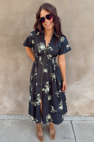 Woman wearing a floral dress standing against a plain wall.