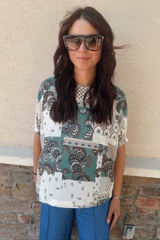 Woman wearing a patterned shirt and sunglasses against a beige wall.