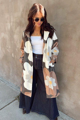 Woman wearing a floral cardigan, white top, and dark pants against a beige wall.