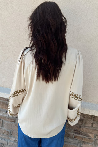 Person wearing a cream-colored blouse with gold embroidery and blue pants against a beige wall.