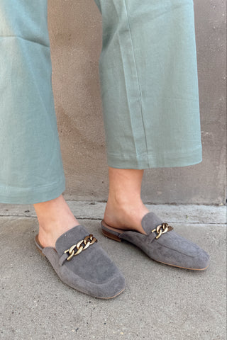 Person wearing gray loafers with gold chains on a concrete surface.