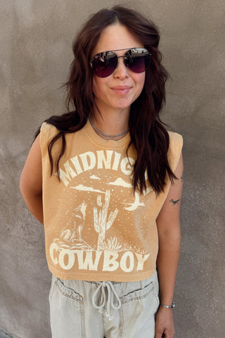 Woman wearing a mustard yellow tank top with graphic design and text, standing against a concrete wall.