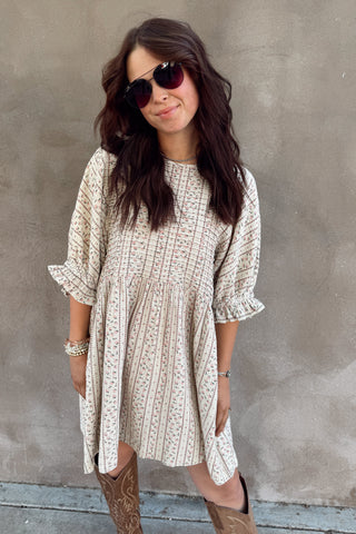 Woman wearing a patterned dress and sunglasses against a gray wall.