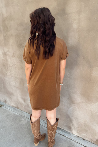 Person wearing a brown dress and cowboy boots standing against a beige wall.