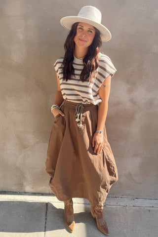 Woman wearing a striped shirt, wide-brimmed hat, and brown skirt against a plain wall.