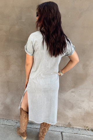 Woman wearing a long light gray dress and brown boots against a beige wall.