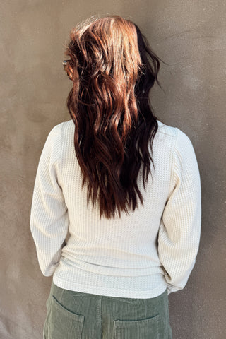 Person with long, wavy hair wearing a white sweater against a brown wall