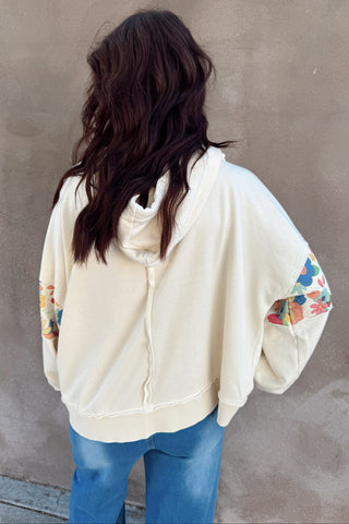 Person wearing a cream-colored hoodie with colorful designs on sleeves against a plain wall.