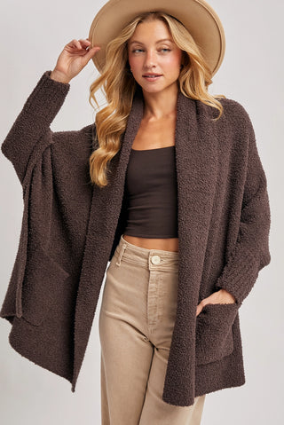 Woman wearing a brown cardigan, black top, and beige pants with a beige hat on a light gray background