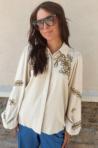 Woman wearing a cream blouse with gold embroidery and sunglasses against a stone wall.