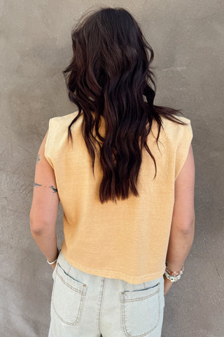 Person wearing a yellow sleeveless top and light blue jeans against a gray wall.