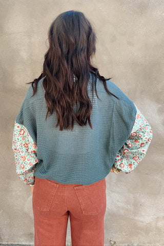 Person wearing a green textured sweater over a floral shirt with red pants, standing against a beige wall.