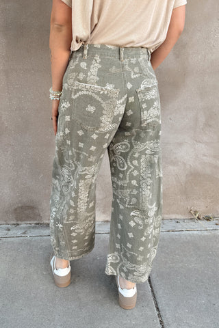 Person wearing patterned green pants and a beige top against a plain wall.