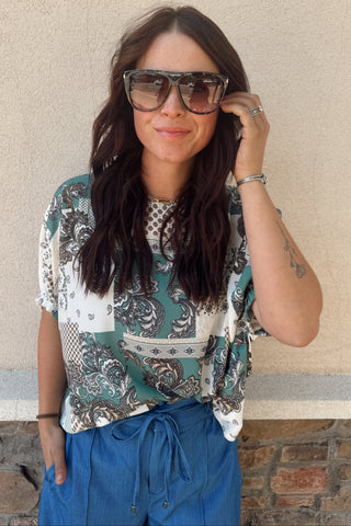 Woman wearing sunglasses and a patterned top against a beige wall.