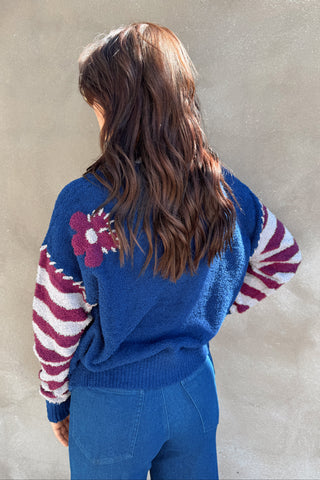 Person wearing a blue sweater with a floral design on the back, standing against a neutral wall.