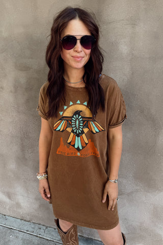 Woman wearing a brown dress with a graphic design, sunglasses, and cowboy boots against a concrete wall.