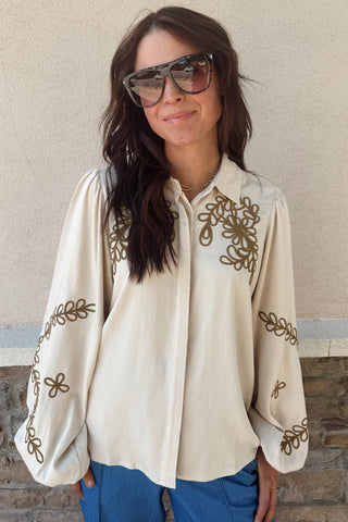 Woman wearing a cream blouse with gold embroidery against a beige wall.