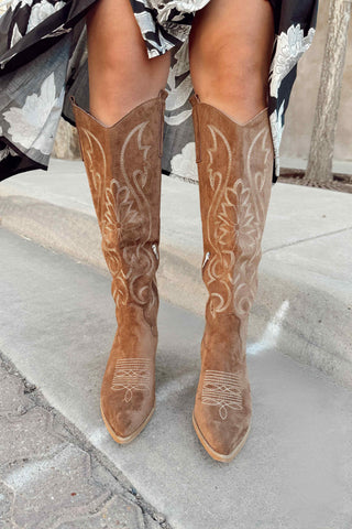 Brown cowboy boots with white embroidery worn by a person sitting on a concrete step.