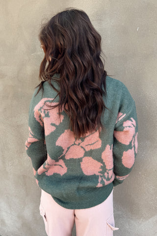 Person wearing a green sweater with pink floral patterns against a beige wall.