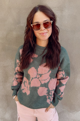 Woman wearing a green sweater with pink floral patterns and pink pants against a gray wall.