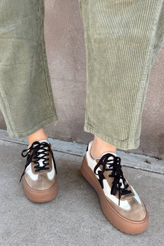 Person wearing brown sneakers with white soles and black laces, paired with green corduroy pants against a neutral background.