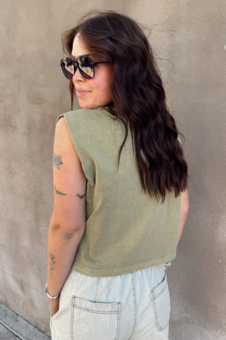 Woman wearing a green sleeveless top and light-colored pants against a plain wall.