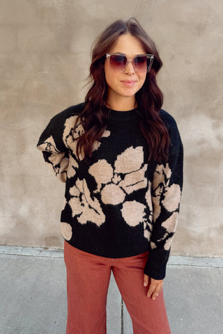 Woman wearing a black sweater with beige floral patterns and pink pants against a beige wall.