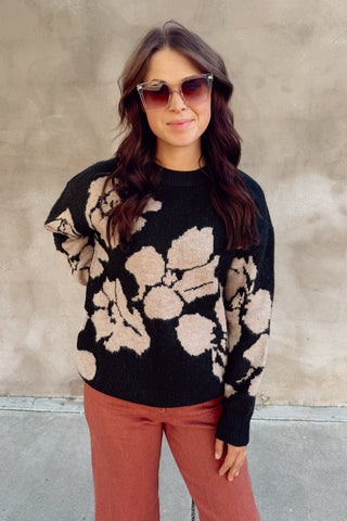Woman wearing a black sweater with beige floral patterns and pink pants against a concrete wall.