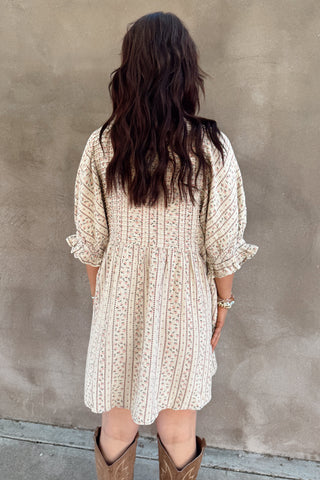 Person wearing a patterned dress and boots standing against a plain wall.