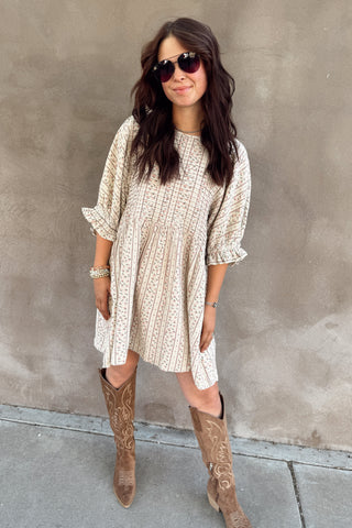 Woman wearing a patterned dress and brown knee-high boots against a gray wall.