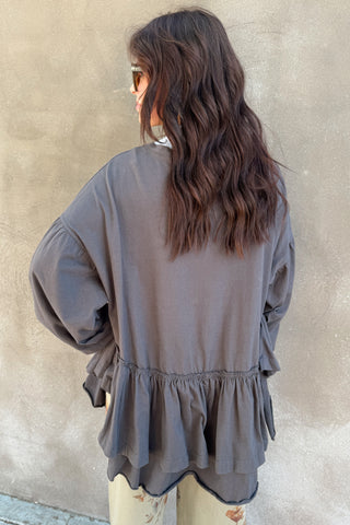 Person wearing a gray long-sleeve top with ruffled details against a beige wall.