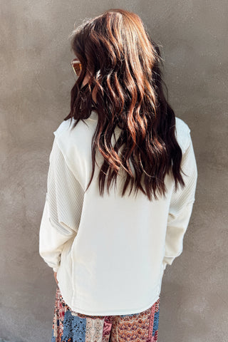 Person with long wavy hair wearing a white sweater against a plain wall