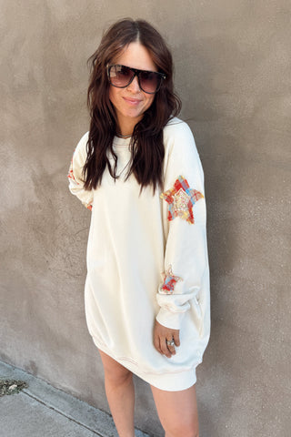 Woman wearing a white sweatshirt dress with colorful designs against a beige wall.
