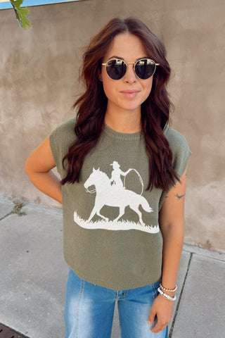 Woman wearing an olive green t-shirt with a horse and rider graphic, standing against a beige wall.