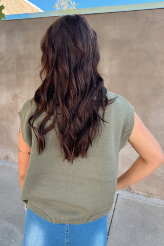 Person wearing a green sleeveless sweater and blue jeans standing against a concrete wall with a clear blue sky.