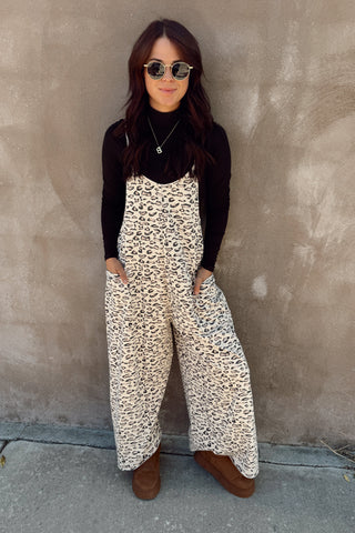 Person wearing a black long-sleeve top and white leopard print jumpsuit against a beige wall.